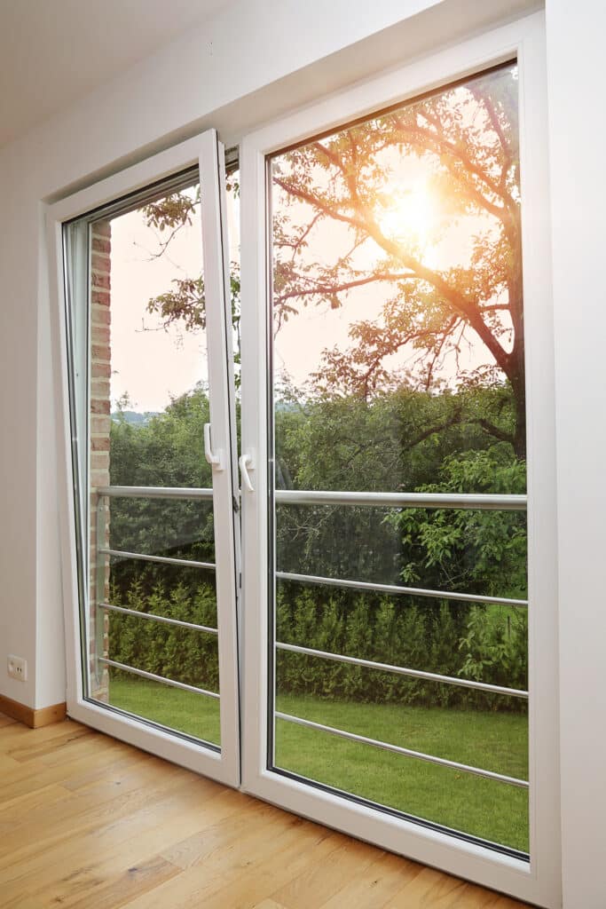 Porte-fenêtre PVC vue sur jardin et soleil couchant Intérieur lumineux avec sol en parquet clair ouvrant sur un jardin verdoyant via une porte-fenêtre oscillo-battante PVC blanche.