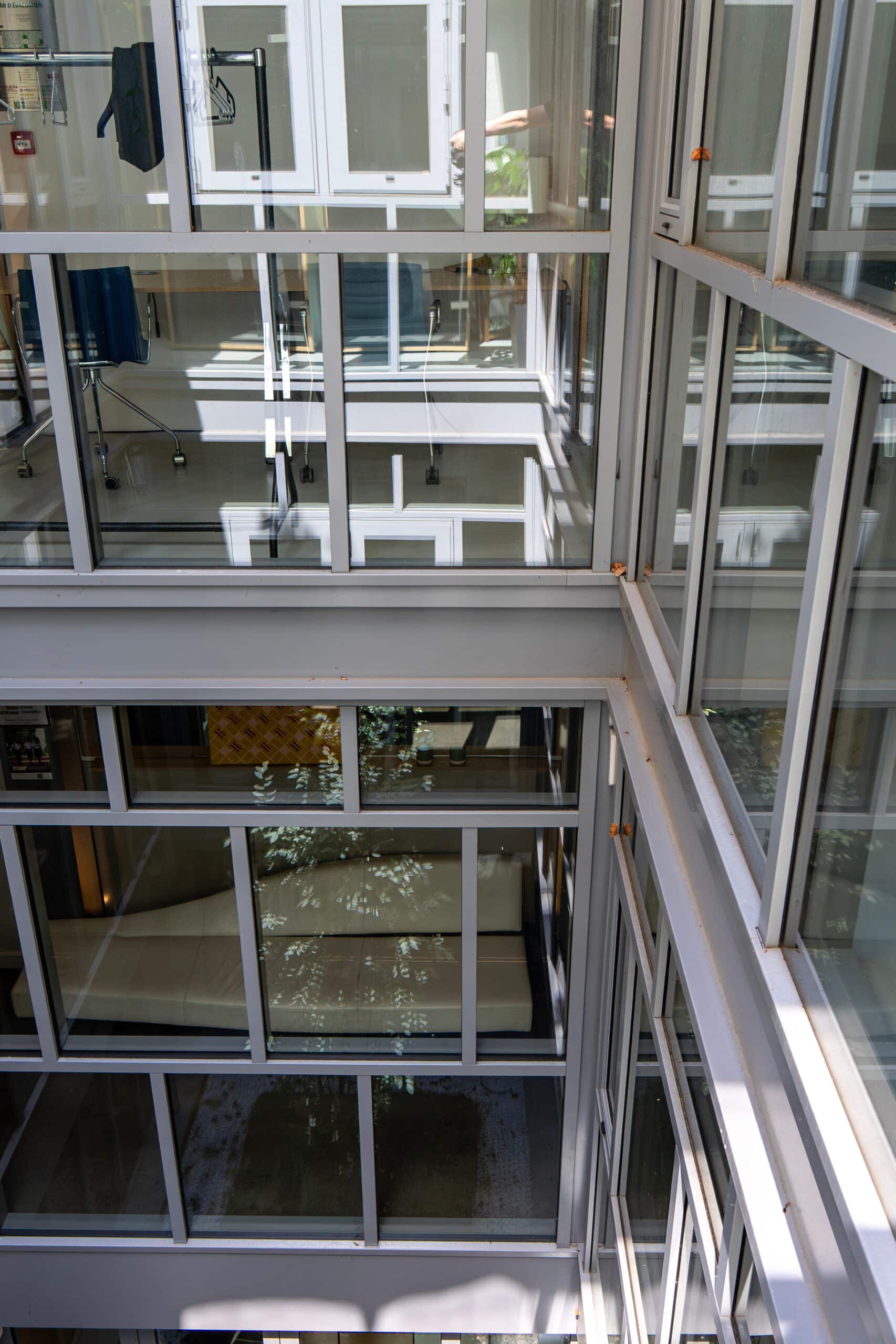Structure moderne d'atrium intérieur à plusieurs niveaux, vue plongeante à travers des cadres de fenêtres gris et blancs, reflets végétaux.