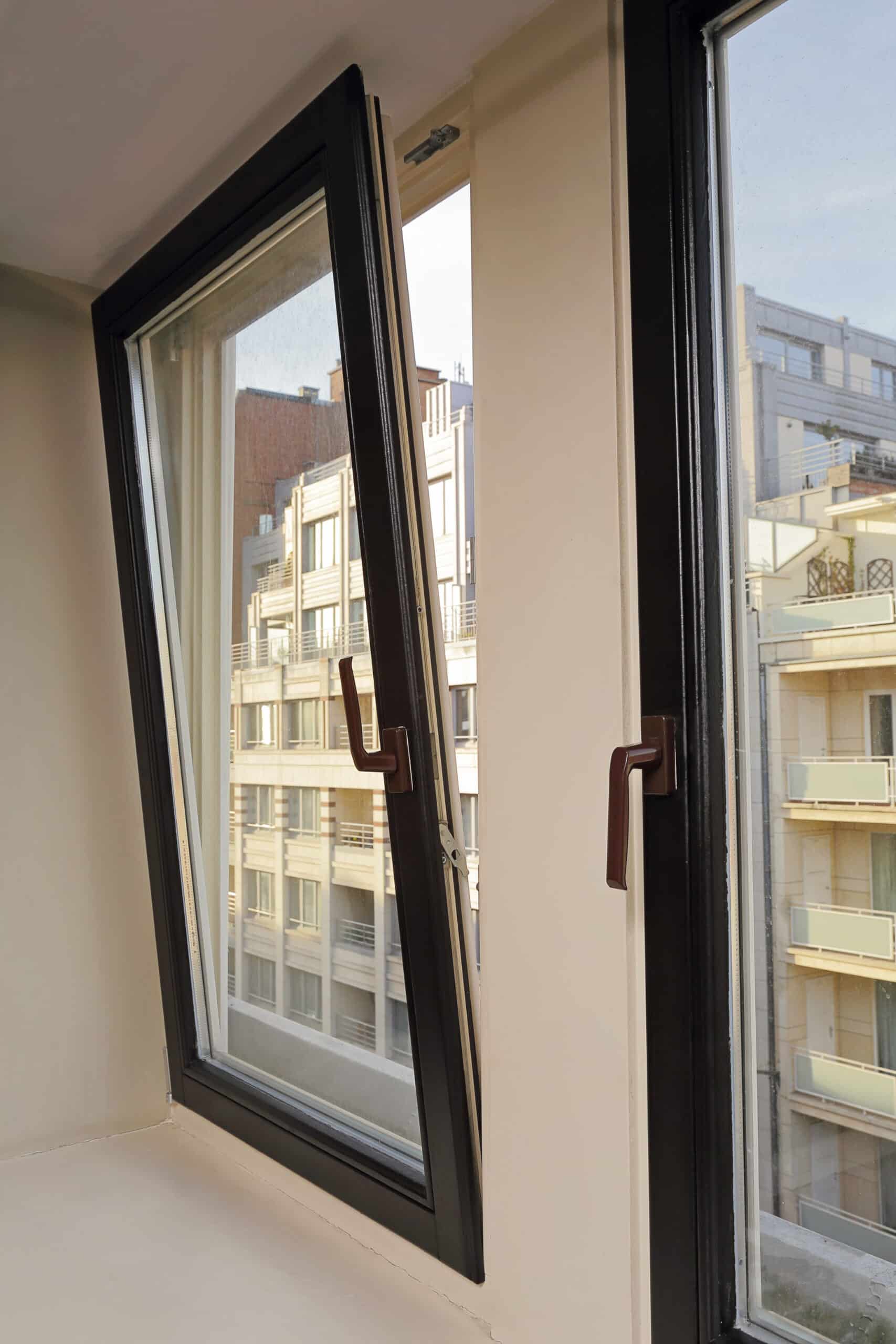 Fenêtre oscillo-battante noire ouverte en inclinaison, vue sur des immeubles urbains clairs et des balcons.