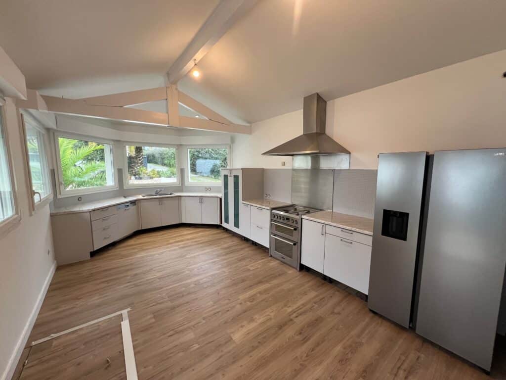 Cuisine Moderne Inox et Lumière : Aménagement Neuf Cuisine moderne avec armoires claires, plan de travail clair, four et hotte en inox, et grand réfrigérateur Samsung. Grandes fenêtres donnent sur la végétation extérieure.